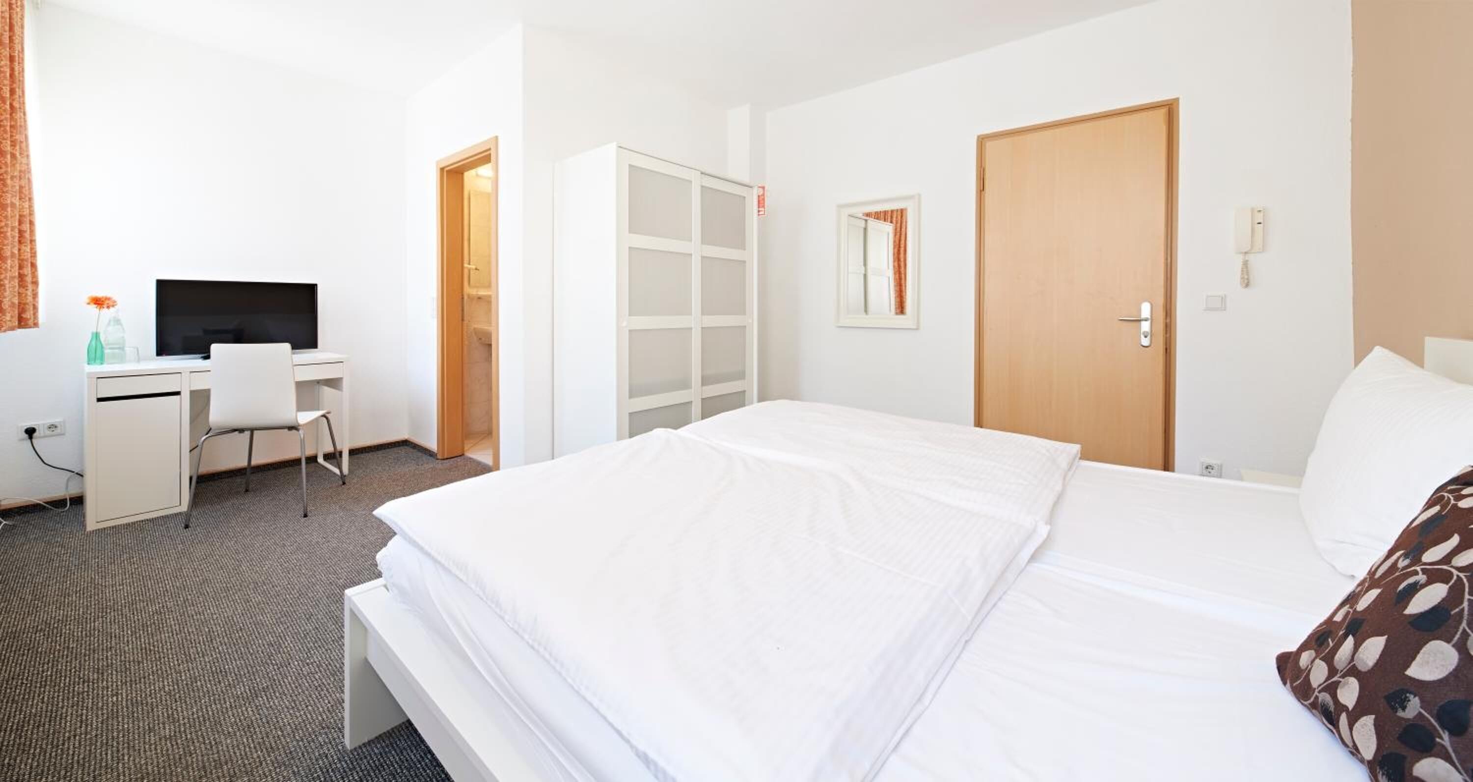 Ein Hotelzimmer mit Bett, Schreibtisch mit Stuhl, Fernseher, Kleiderschrank und einer Tür.