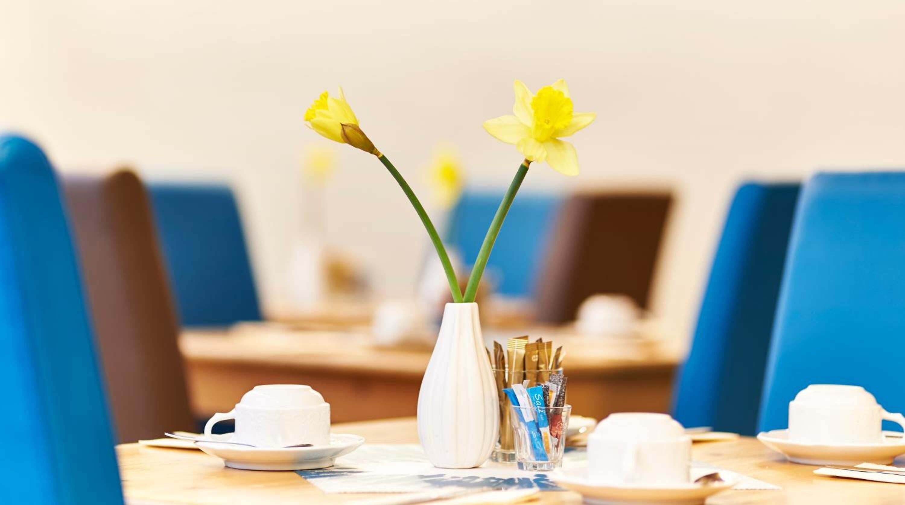A dining table with a white vase containing yellow flowers, a glass of water, and a white cup and saucer.