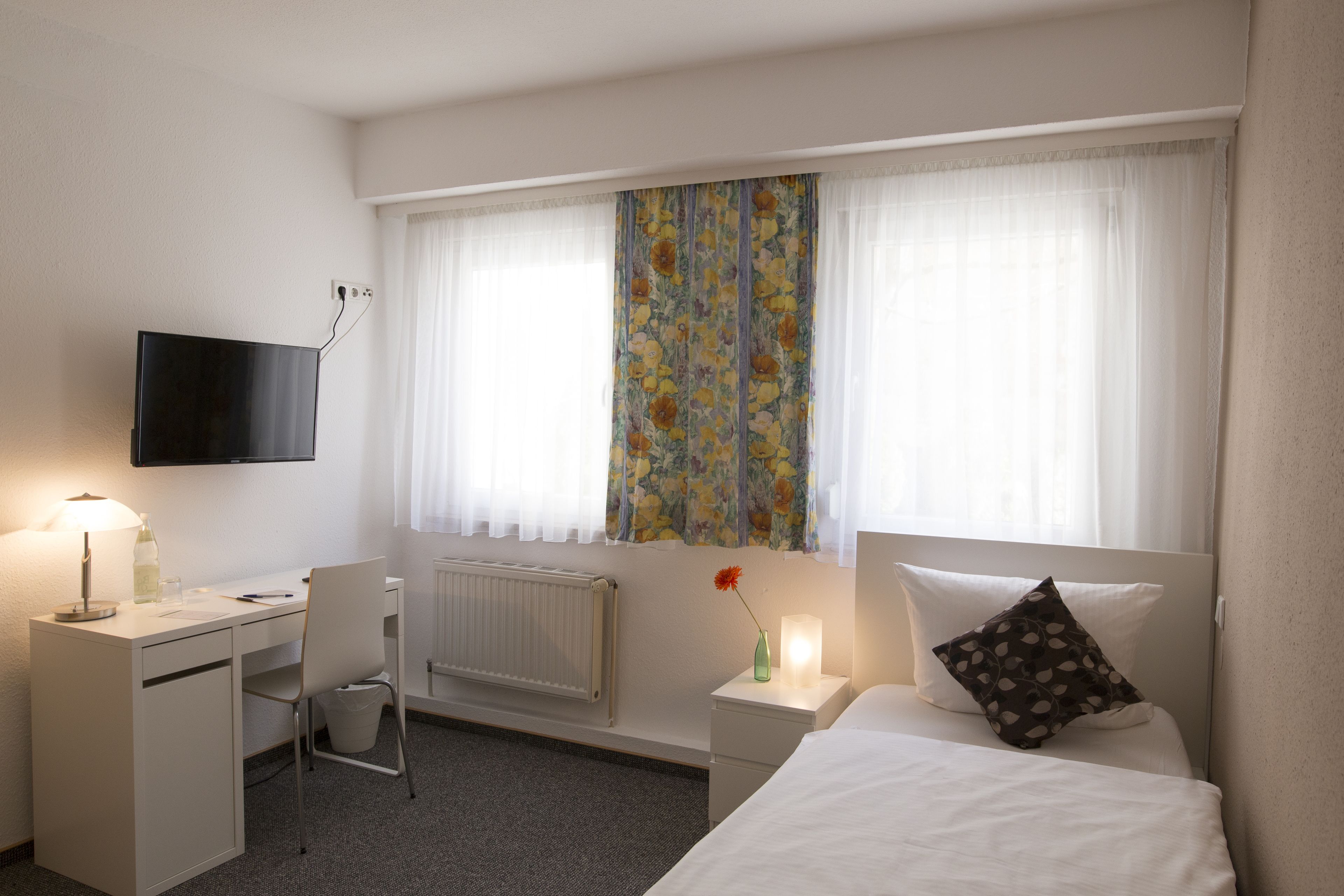 Hotelkamer met een bed, bureau met stoel, televisie aan de muur, radiator en raam met bloemen gordijnen.