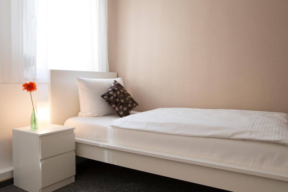 A minimalist bedroom with a single bed, a nightstand, a vase with a flower, and a window with sheer curtains.