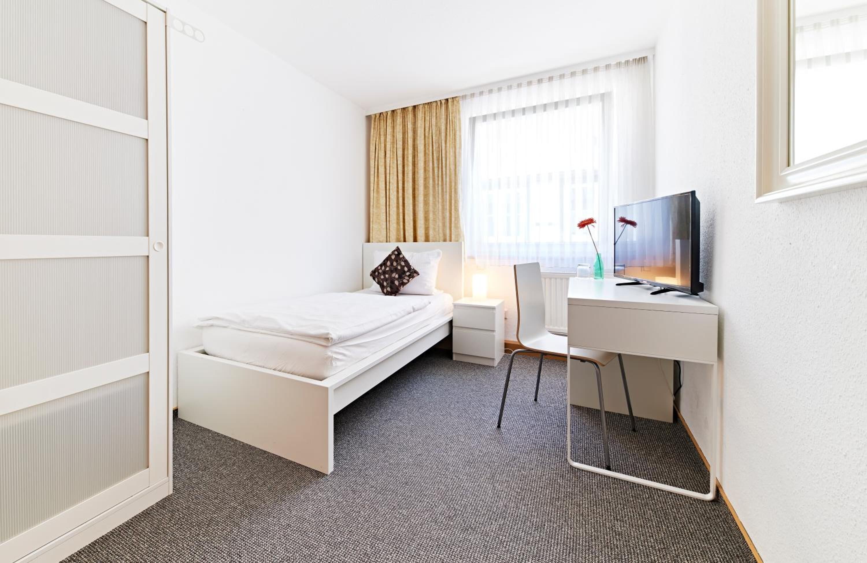 A compact hotel room with a bed, a desk with a chair, a television, and a window with curtains.