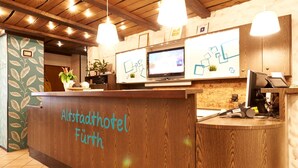 A reception desk with a wooden finish and the name "Altstadthotel Fürth" written on it. There are two monitors on the desk, a plant, and a small shelf with items. The ceiling has wooden beams and hanging lights. - Altstadthotel Fürth (Fuerth)