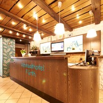 Un mostrador de recepción con detalles de madera y el nombre "Altstadthotel Fürth" escrito en él. Hay dos monitores en el mostrador, una planta y una pequeña estantería con objetos. El techo tiene vigas de madera y luces colgantes.