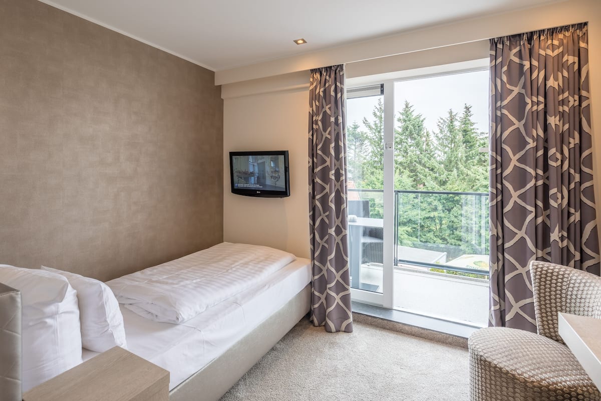 Ein Hotelzimmer mit Bett, einem an der Wand montierten Fernseher, einem Balkon mit Vorhängen und einer Sitzecke mit einem Sessel.