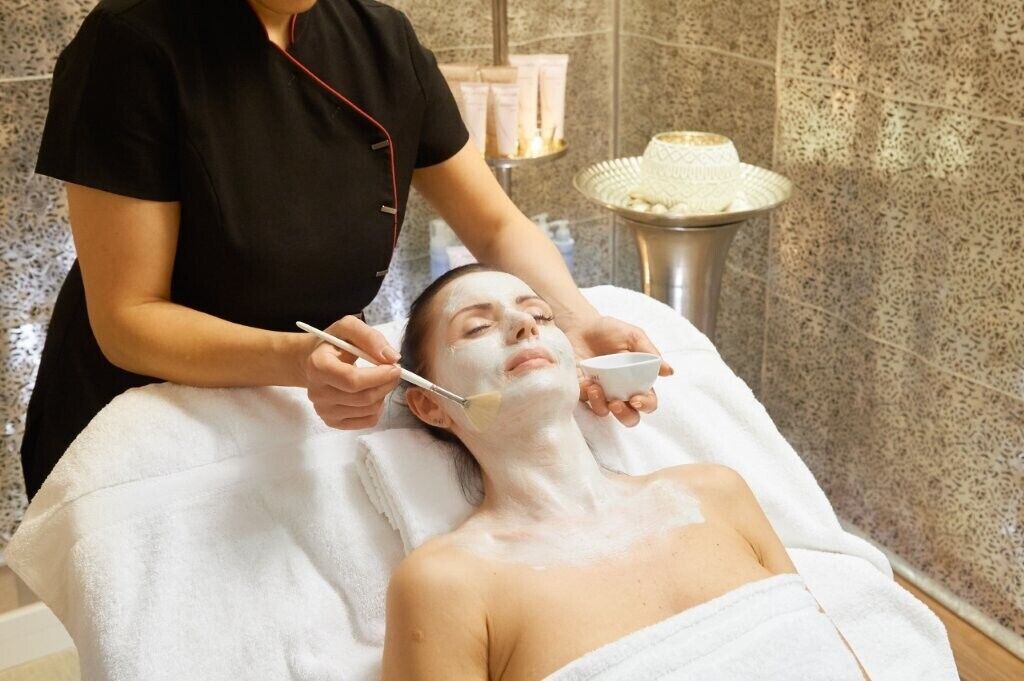 A person receiving a facial treatment in a spa setting with a tiled wall and a small table with products.