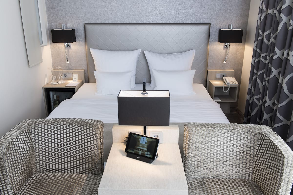 Ein Hotelzimmer mit einem großen Bett, zwei Nachttischen mit Lampen, einem Nachttischtelefon, einem kleinen Tisch mit einem Tablet und zwei gemusterten Sesseln.