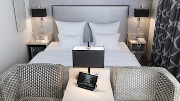 Een hotelkamer met een groot bed, twee nachtkastjes met lampen, een telefoon op het nachtkastje, een klein tafeltje met een tablet, en twee gestreept beklede fauteuils.