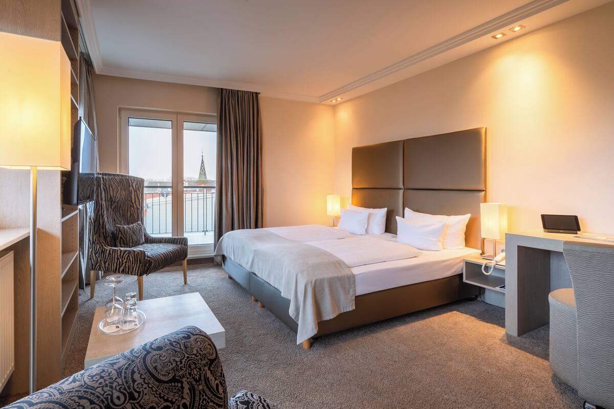 Ein Hotelzimmer mit einem großen Bett, zwei Nachttischen, einem Sessel, einem Schreibtisch und Blick auf ein Gebäude durch ein Fenster.