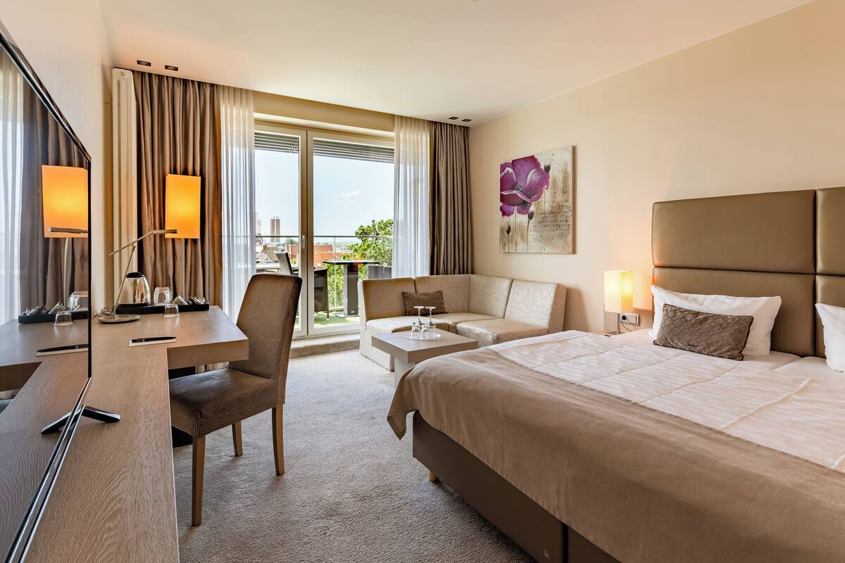 Ein Hotelzimmer mit einem großen Bett, einem Schreibtisch mit Stuhl, einer Couch, einem kleinen Tisch und einem Balkon mit Vorhängen.