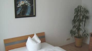 Ein ordentlich bezogenes Bett mit weißer Bettwäsche und Kissen, ein hölzerner Kopfteil, ein gerahmtes Kunstwerk darüber und eine Zimmerpflanze rechts.