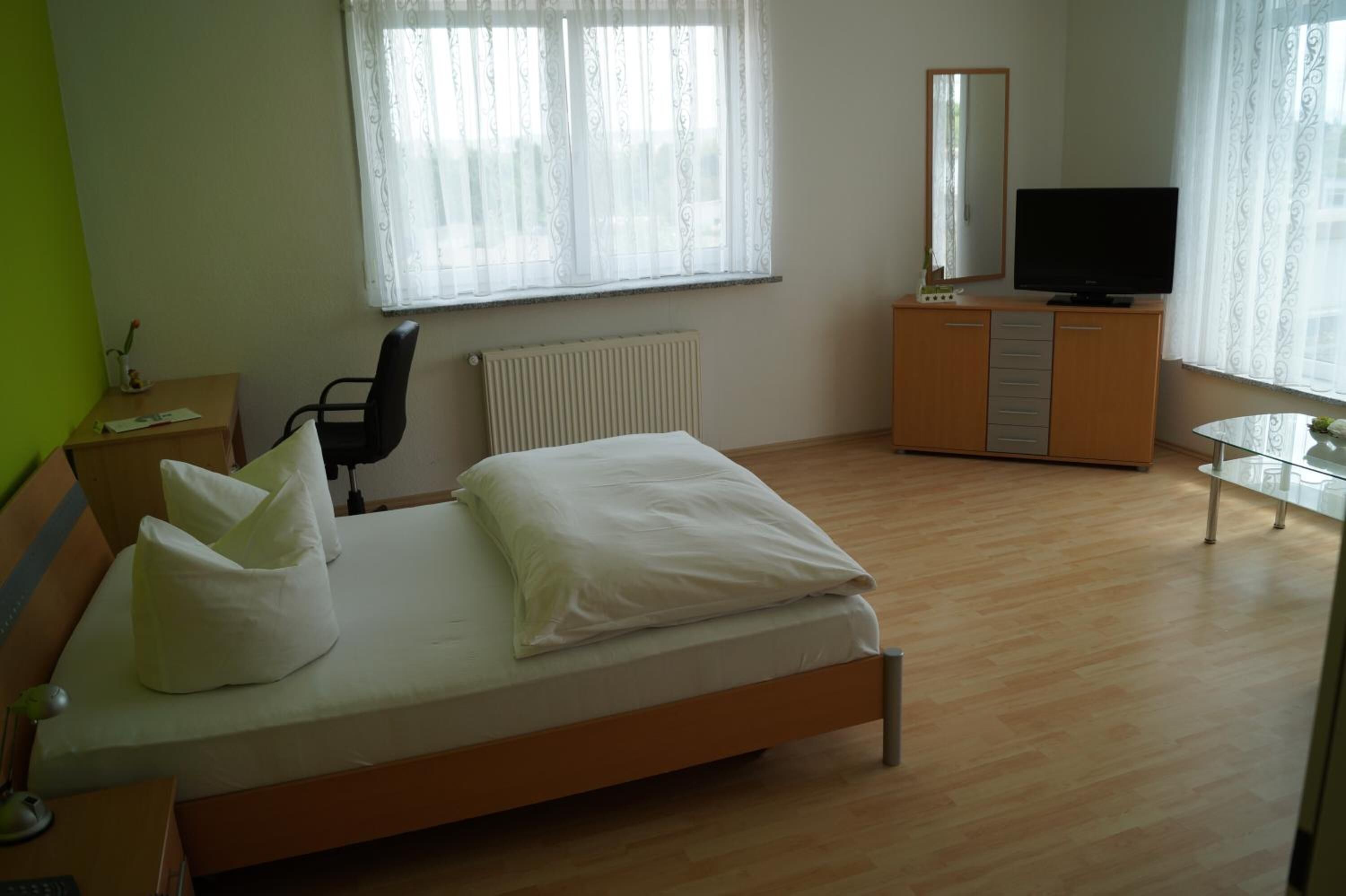 Ein Schlafzimmer mit einem Bett, einem Schreibtisch, einem Stuhl, einer Kommode, einem Fernseher und einem Fenster mit Vorhängen.