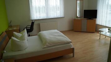 Ein Schlafzimmer mit einem Bett, einem Schreibtisch, einem Stuhl, einer Kommode, einem Fernseher und einem Fenster mit Vorhängen.