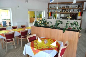 Un restaurante con mesas y sillas de madera, una zona de bar con estanterías llenas de botellas y plantas en macetas.