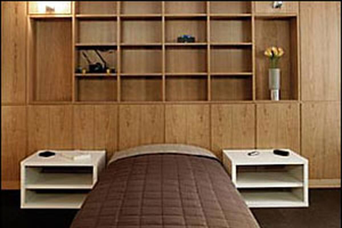 A single bed with a brown quilt, wooden headboard, and footboard, flanked by two white bedside tables with lamps. A wooden shelving unit with a vase of flowers is above the bed.