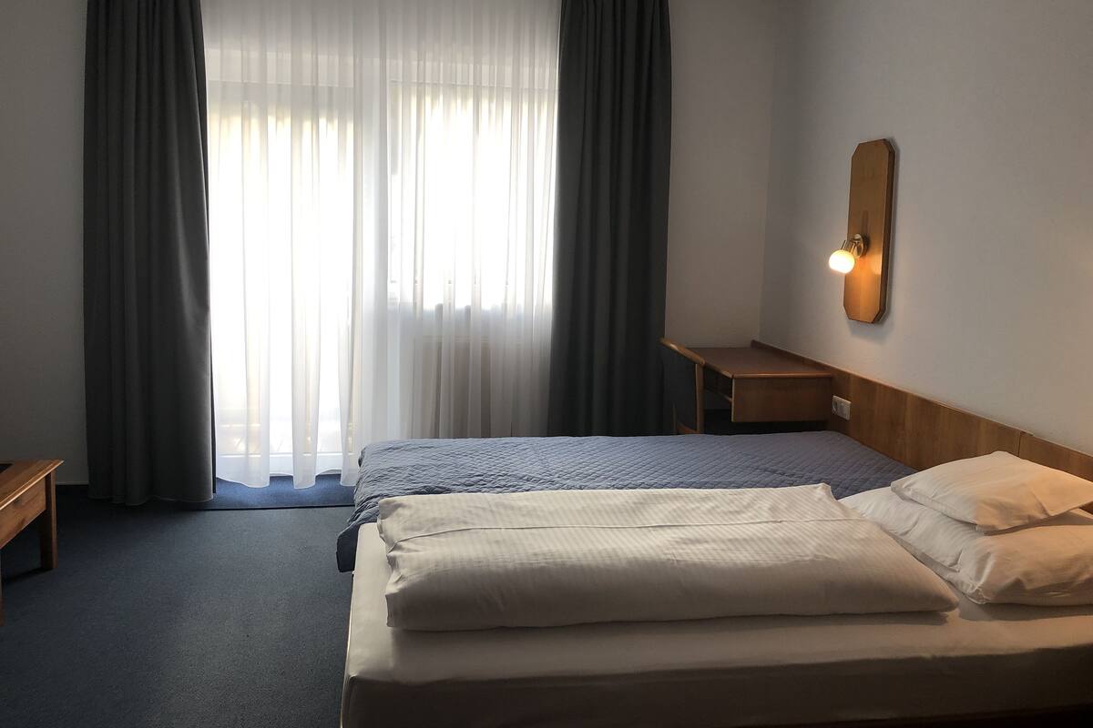 Une chambre d’hôtel avec deux lits, une table de chevet en bois, une lampe fixée au mur et une grande fenêtre ornée de rideaux légers.