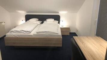 Una cama doble con ropa blanca, dos mesitas de noche con lámparas, un escritorio de madera y una ventana con persianas.