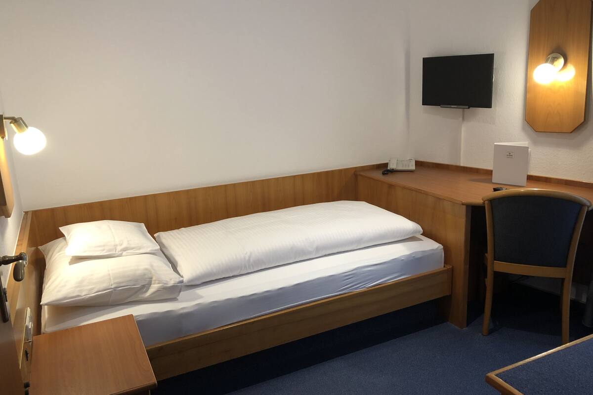 Un lit simple avec une tête de lit en bois, un bureau avec une chaise, un téléviseur à écran plat et une table de chevet avec une lampe.