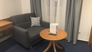 Une chambre d’hôtel avec un canapé gris, une table d’appoint en bois, une petite table ronde sur laquelle se trouve un appareil blanc et une télécommande, et un tapis bleu.