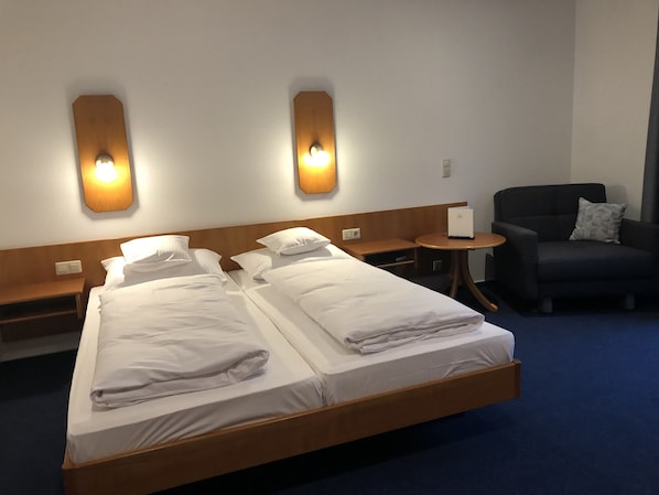 A hotel room with two single beds, a small round table, a chair, and wall-mounted lighting.