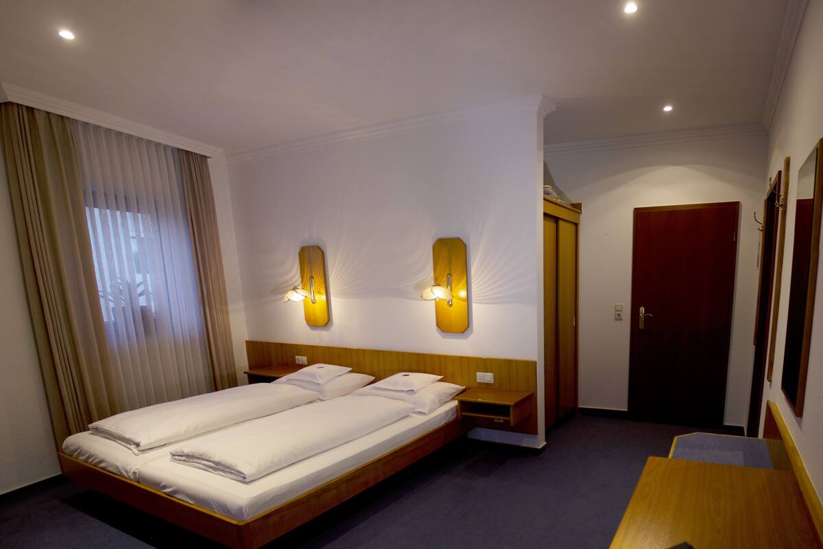 Une chambre d’hôtel avec un lit double, deux lampes fixées au mur, une table de chevet en bois, un bureau et une porte donnant sur une autre pièce.