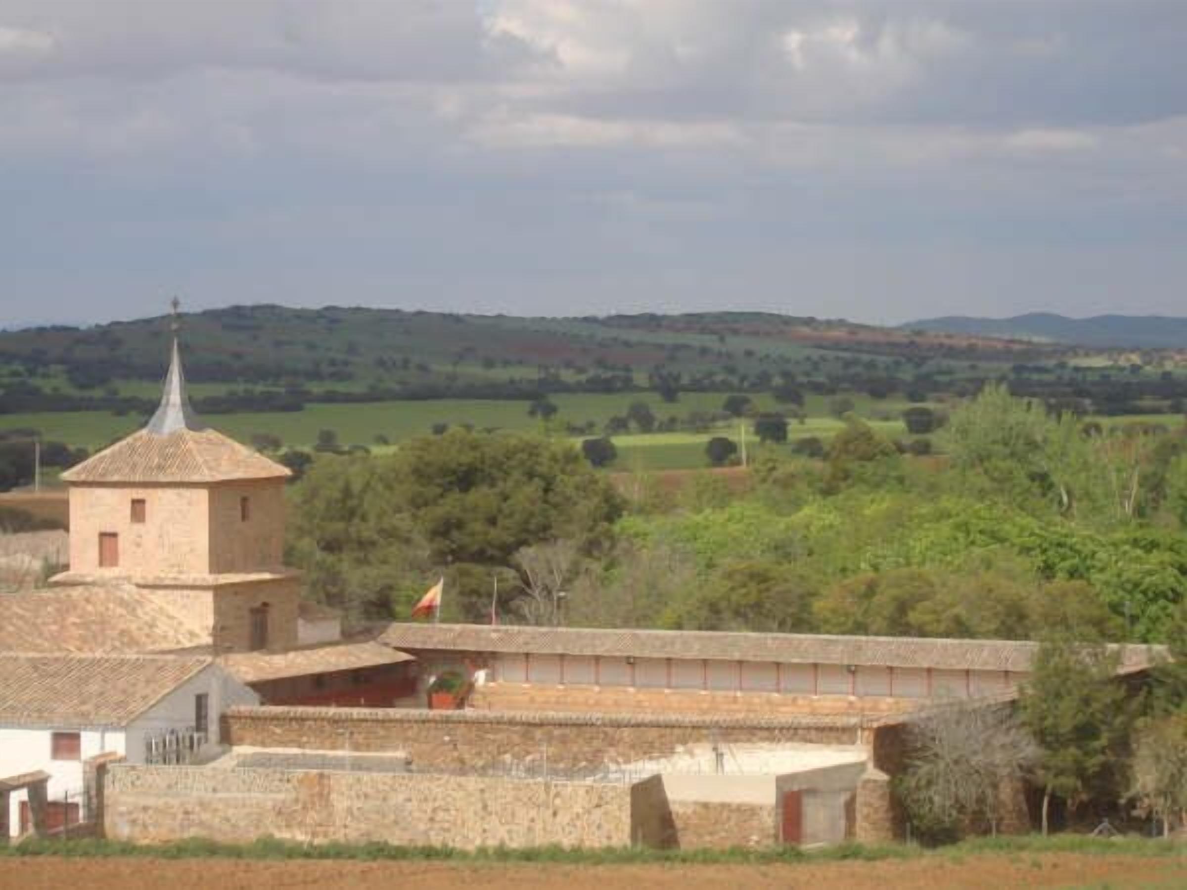 Um edifício rústico com uma pequena torre, rodeado por campos e árvores.