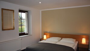 A bedroom with a bed, two bedside lamps, a window with a view, and a radiator. - Landhotel Lindenschänke (Dresden)