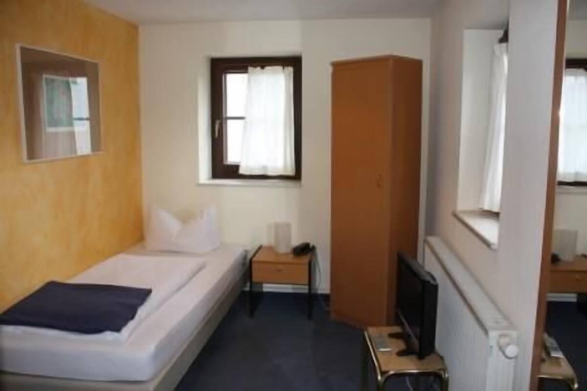 A small, simple room with a bed, a nightstand, a television, and a wardrobe.