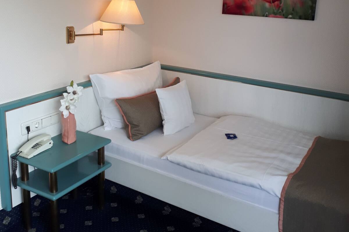 Habitación de hotel ordenada con una cama individual, una mesita de noche con lámpara, un escritorio pequeño con teléfono y un cuadro en la pared.