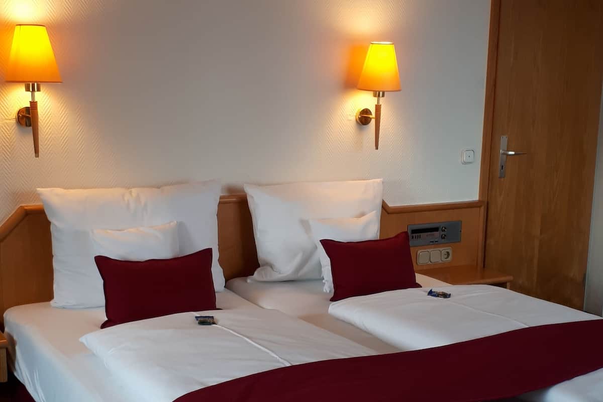 Una cama doble con ropa de cama blanca y roja, dos lámparas fijadas a la pared y una puerta de madera.