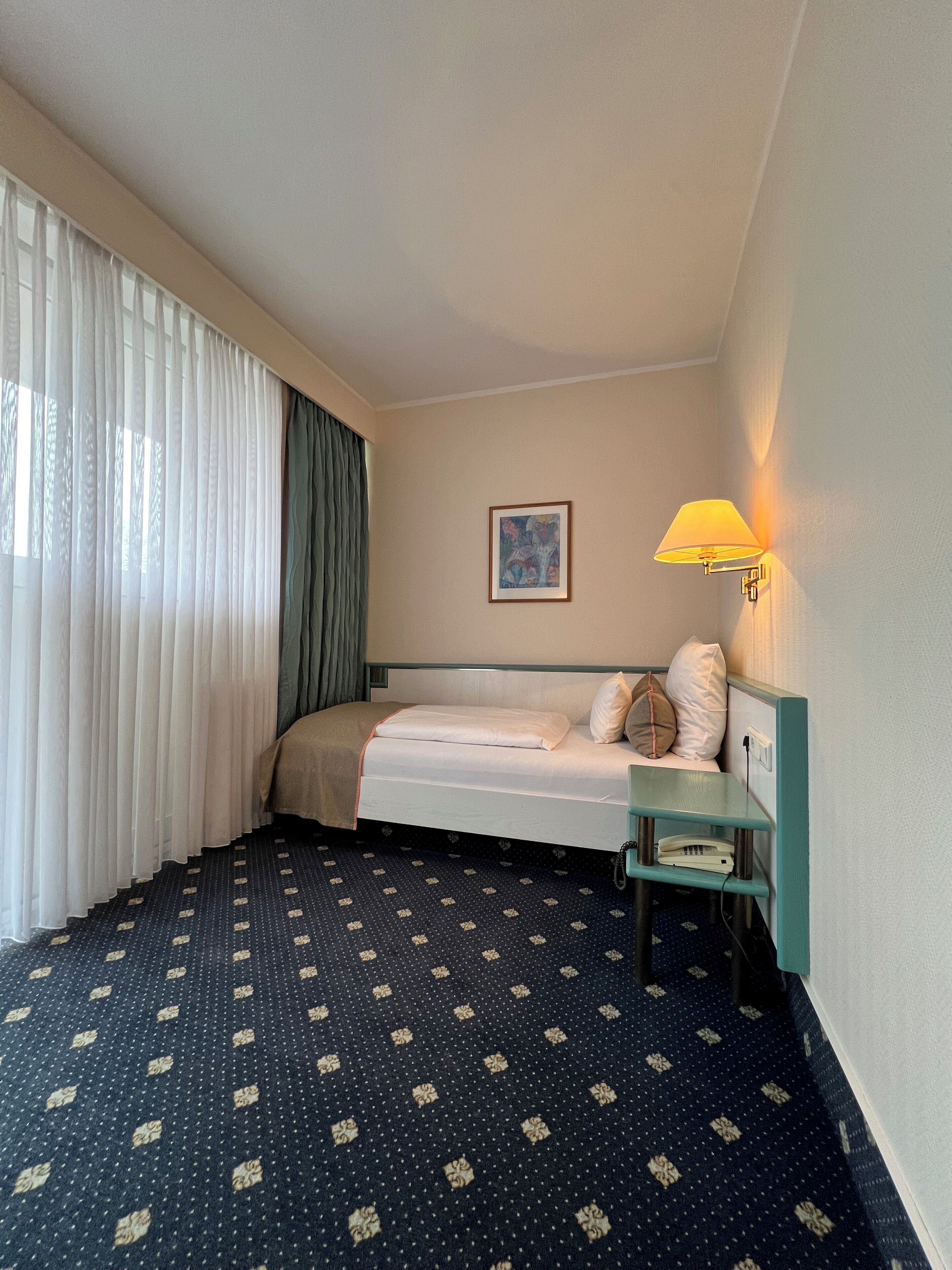 Habitación de hotel con cama, mesita de noche con lámpara, un cuadro en la pared y alfombra estampada.
