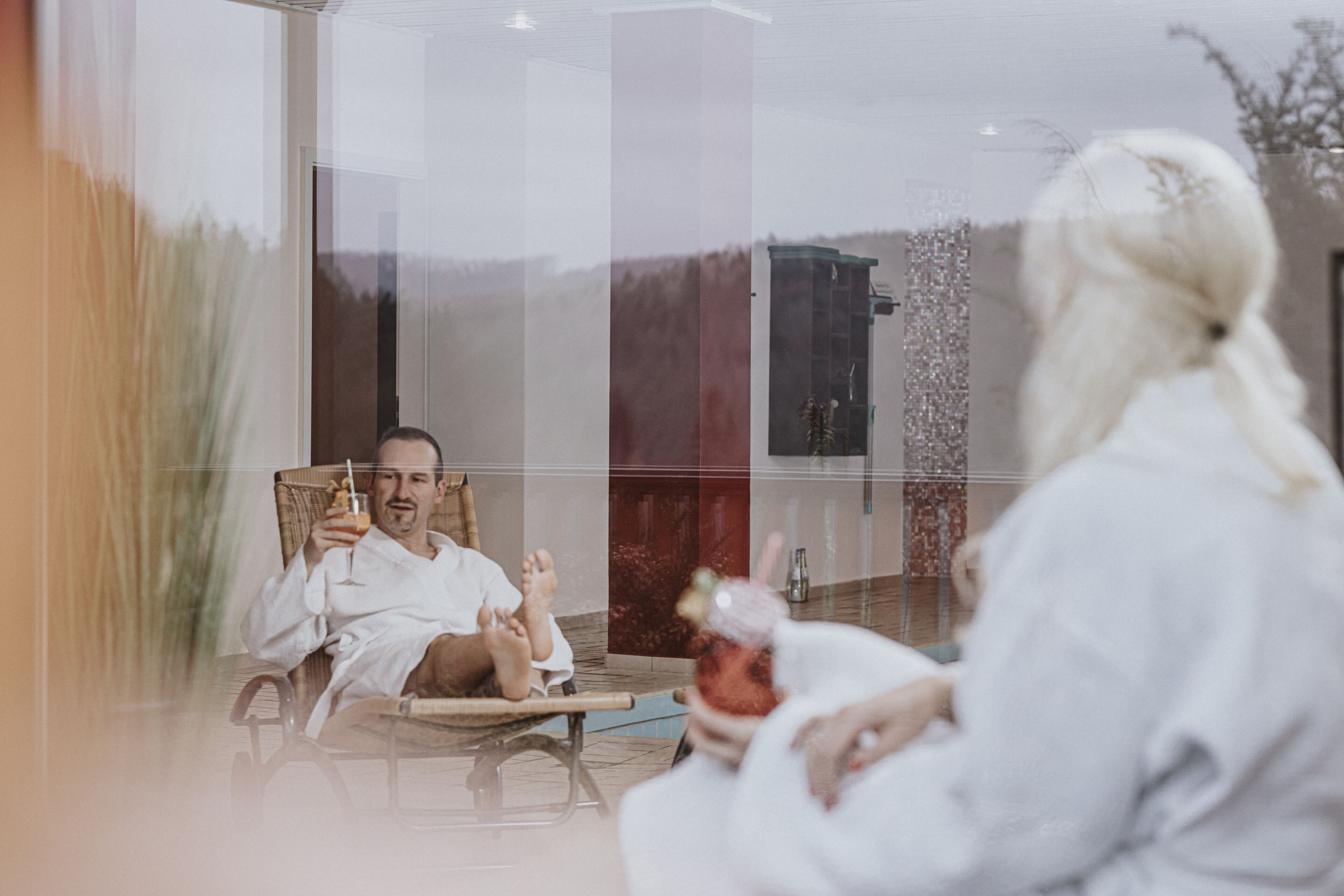 A person in a white robe sitting in a chair with a drink, facing a reflection in a glass window.