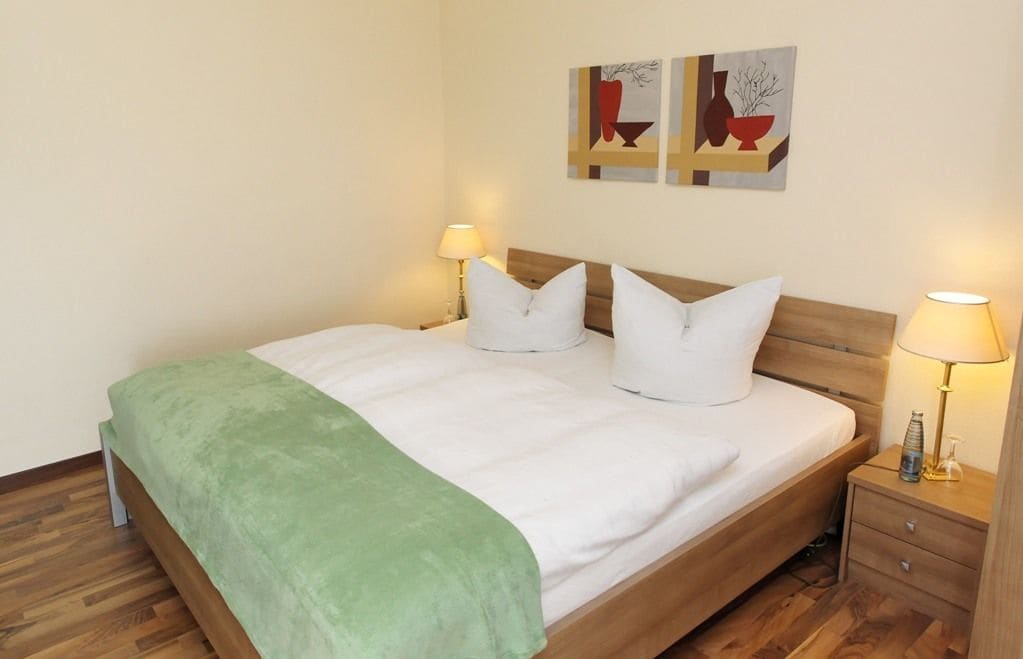 Una cama bien hecha con ropa de cama blanca y una manta verde, flanqueada por dos mesitas de noche con lámparas, en una habitación con suelo de madera y decoración minimalista.