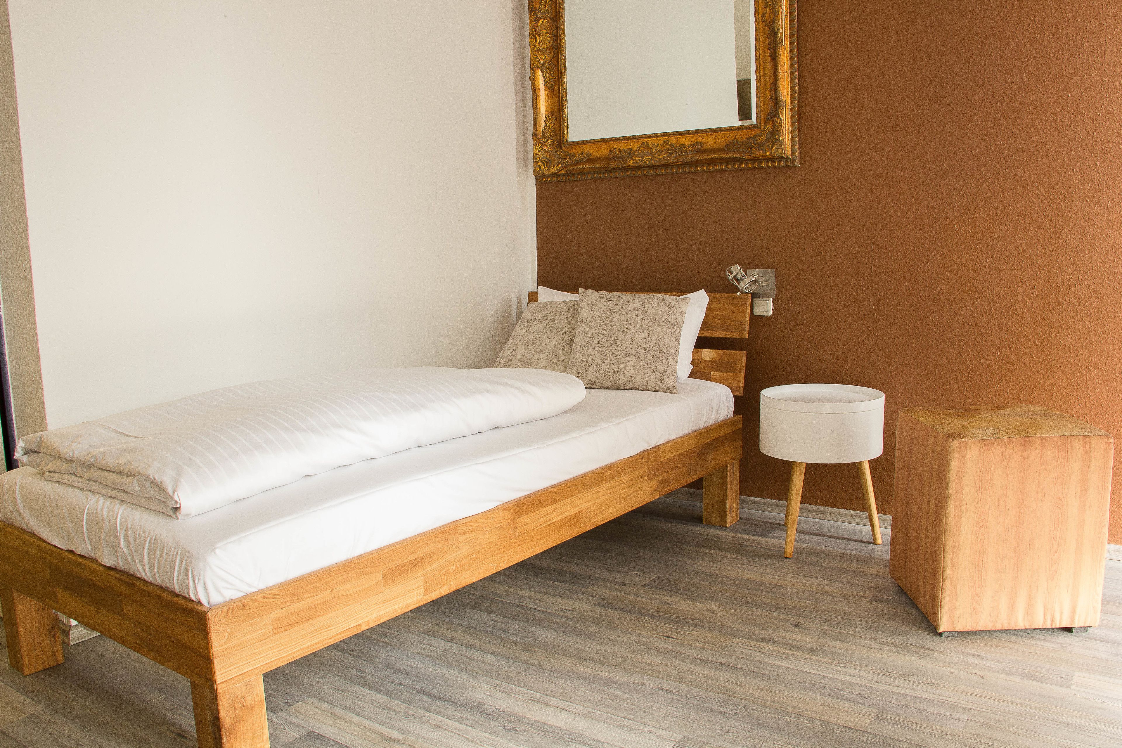 A single bed with a wooden frame, a bedside table, a round nightstand, and a mirror on the wall.