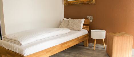 A single bed with a wooden frame, a bedside table, a round nightstand, and a mirror on the wall.