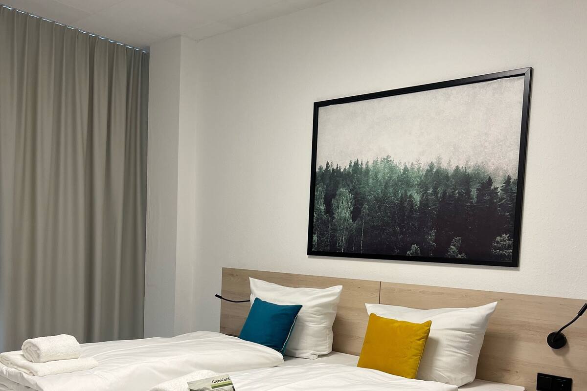 Ett modernt hotellrum med en stor säng, en sänggavel i ljus träimitation, ett nattduksbord med en lampa och en inramad tavla med ett skogsmotiv på väggen.