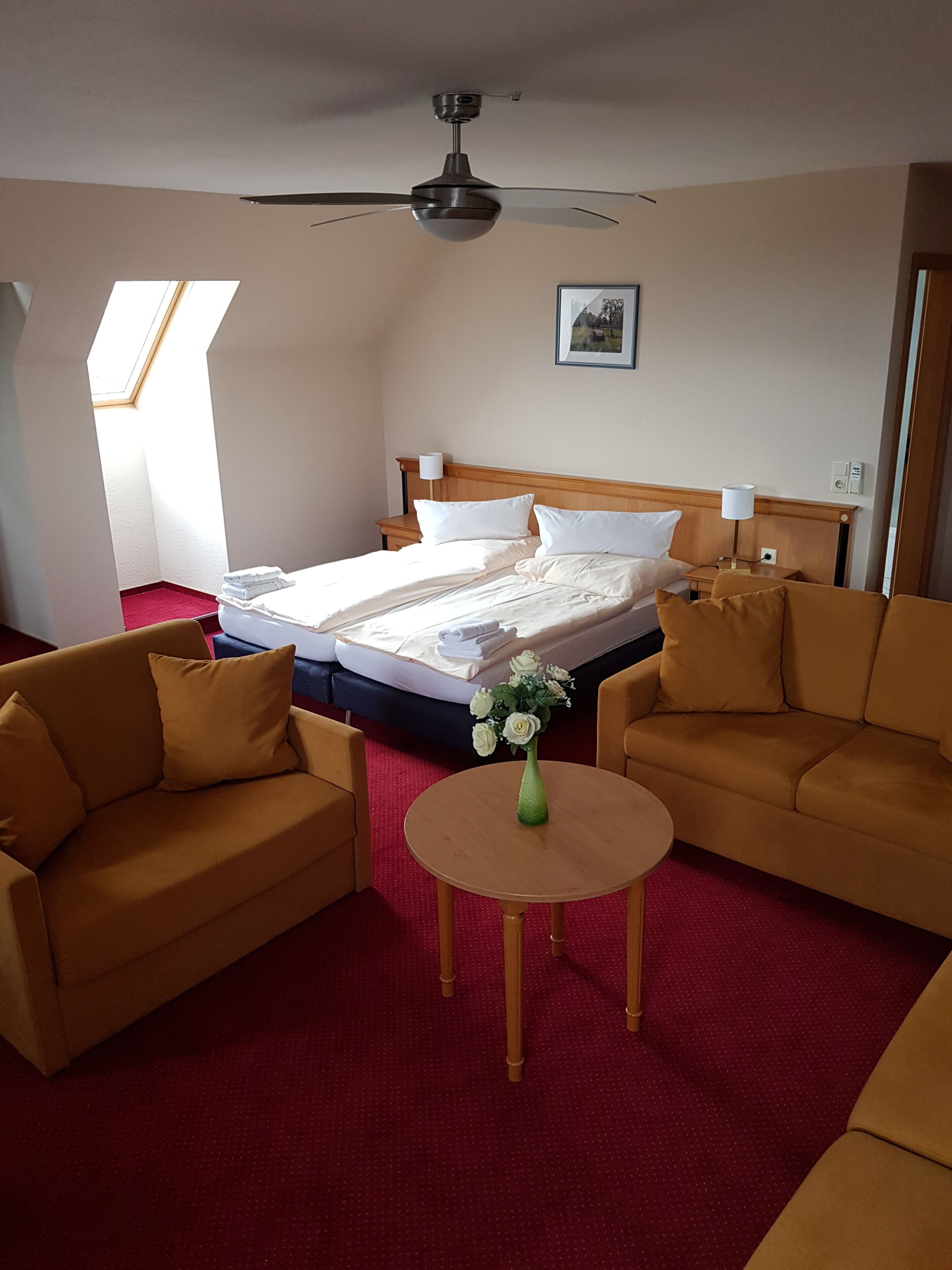 A hotel room with a bed, two sofas, a small table, a ceiling fan, a picture on the wall, and a skylight.