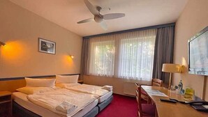 A hotel room with two beds, a desk, a chair, a ceiling fan, a window with curtains, and a wall-mounted TV. - Hotel Radduscher Hafen (Vetschau/Spreewald)