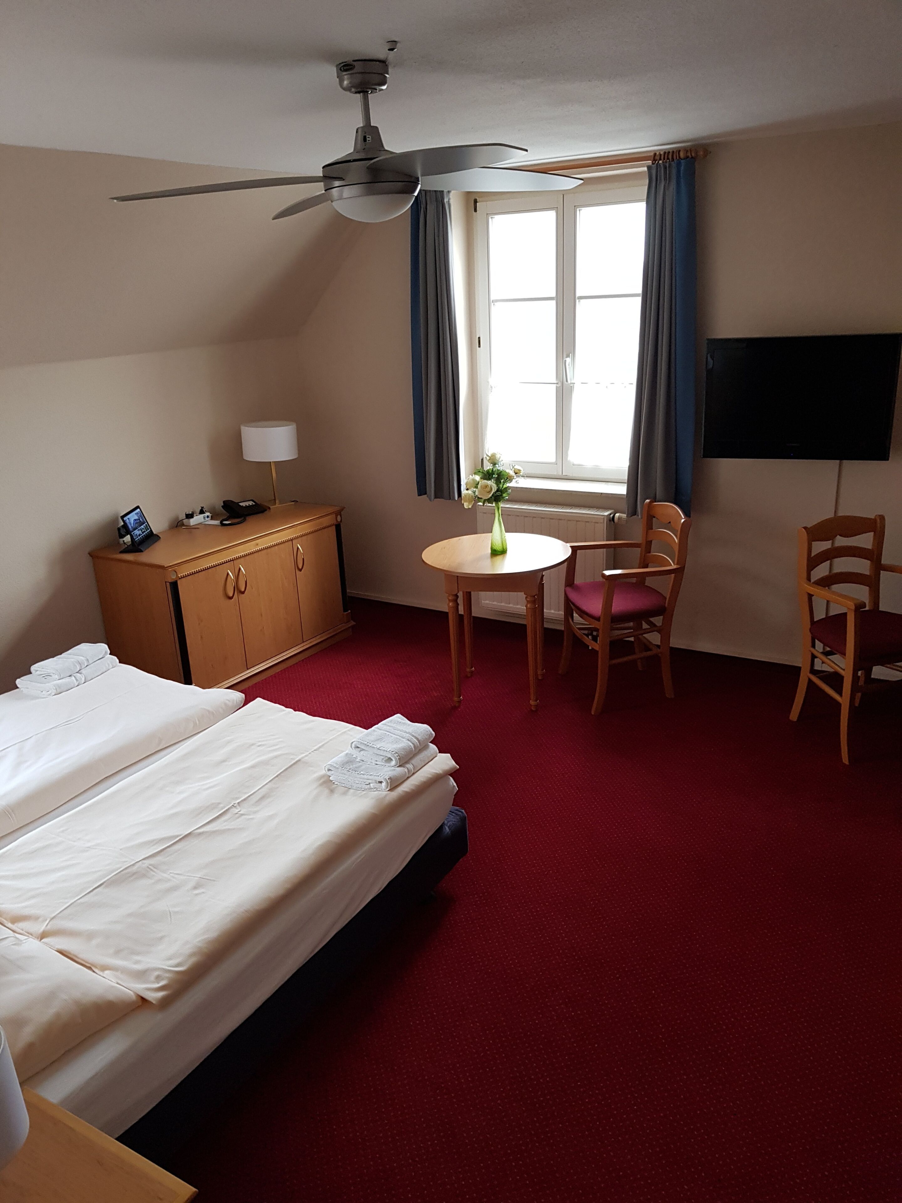 Quarto de hotel com cama, escrivaninha, cadeira, televisão e ventilador de teto.