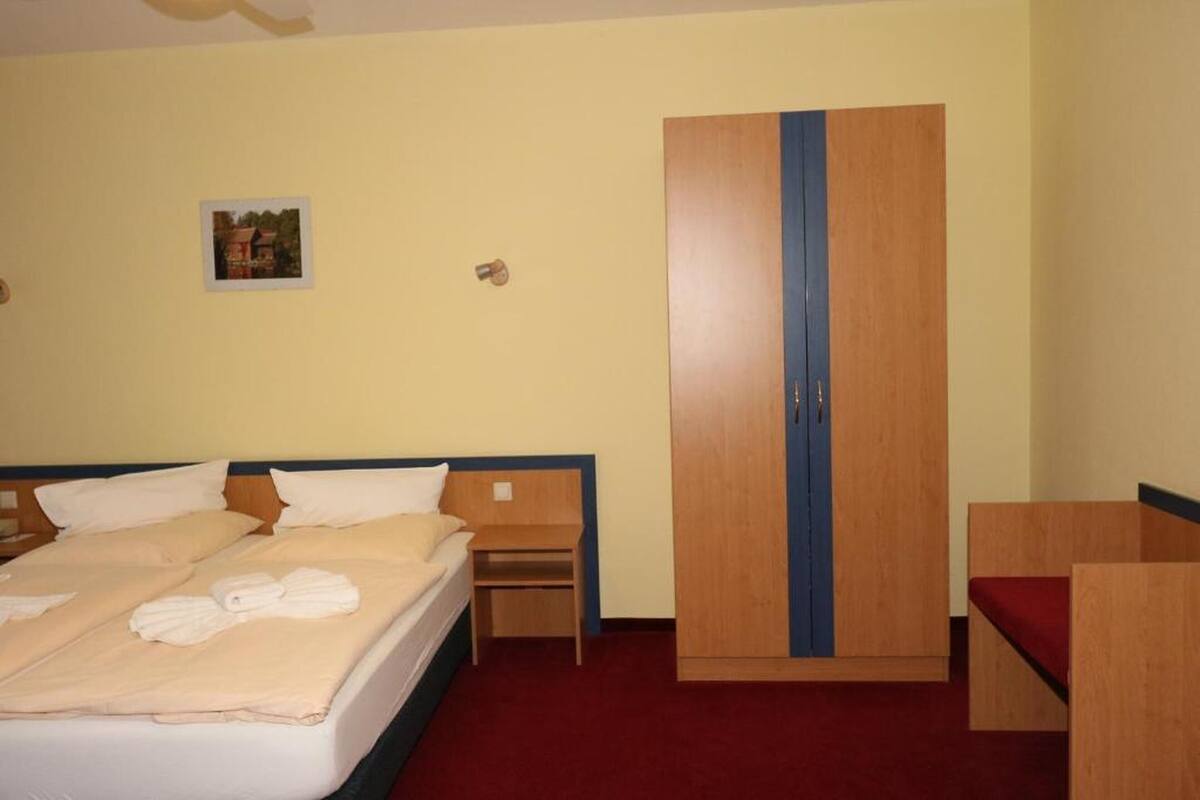 A hotel room with a bed, a wardrobe, a small bedside table, and a red carpet.