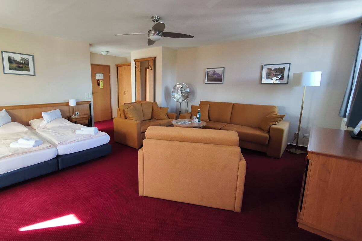 A hotel room with a bed, two sofas, a coffee table, a fan, and a desk.