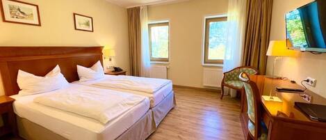 A hotel room with a double bed, a desk with a chair, a television, and two windows with curtains.