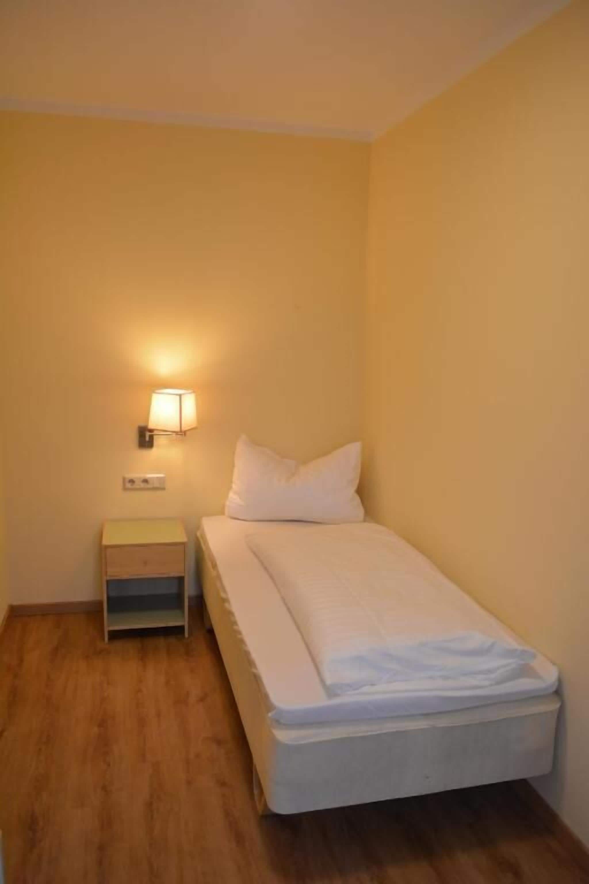 Una habitación pequeña con cama individual, suelo de madera, una mesita de noche con lámpara y pared lisa.