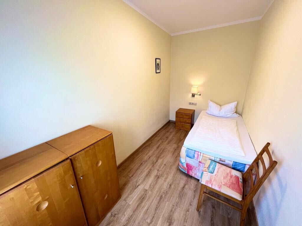 A small, neatly arranged bedroom with a single bed, a wooden wardrobe, a nightstand, and a wall-mounted light.