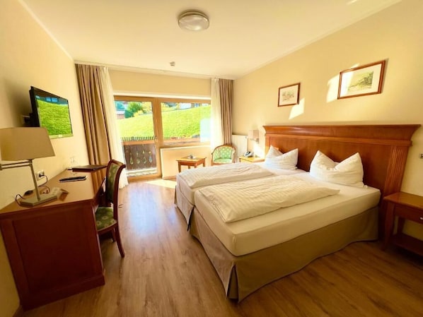 A hotel room with two beds, a desk, a chair, a TV, and a view of a garden through a window.