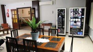 A dining area with a wooden table and chairs, a potted plant, and two beverage vending machines. - Pensión Terminal (Málaga)