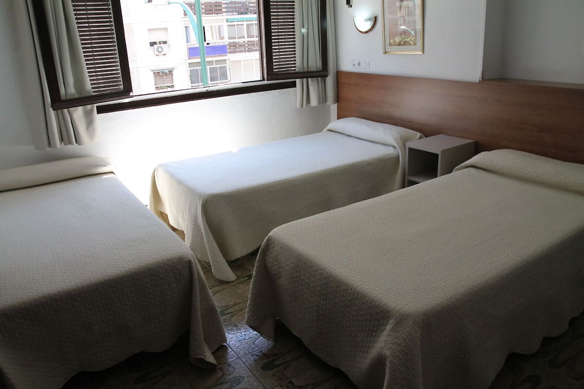 Habitación de hotel con dos camas individuales, cabecero de madera, mesita de noche y ventana con persianas.