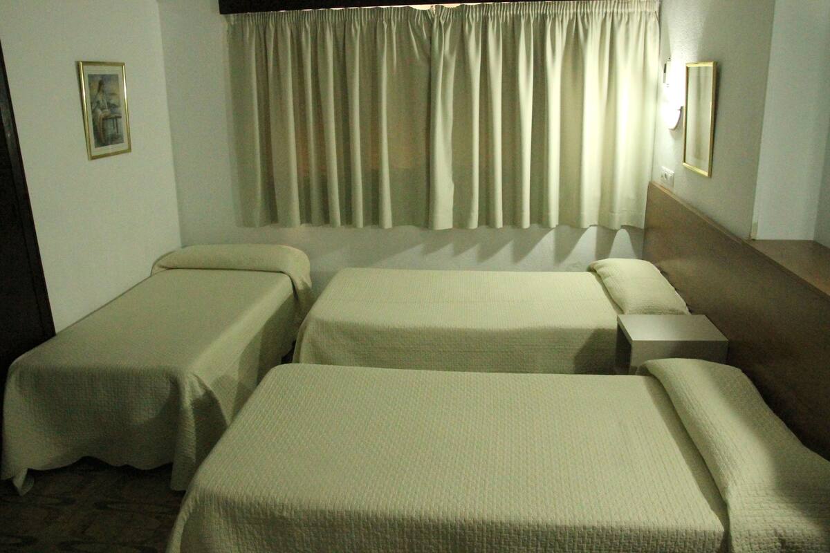 Habitación de hotel con dos camas individuales, una mesita de noche pequeña y un ventanal grande con cortinas.