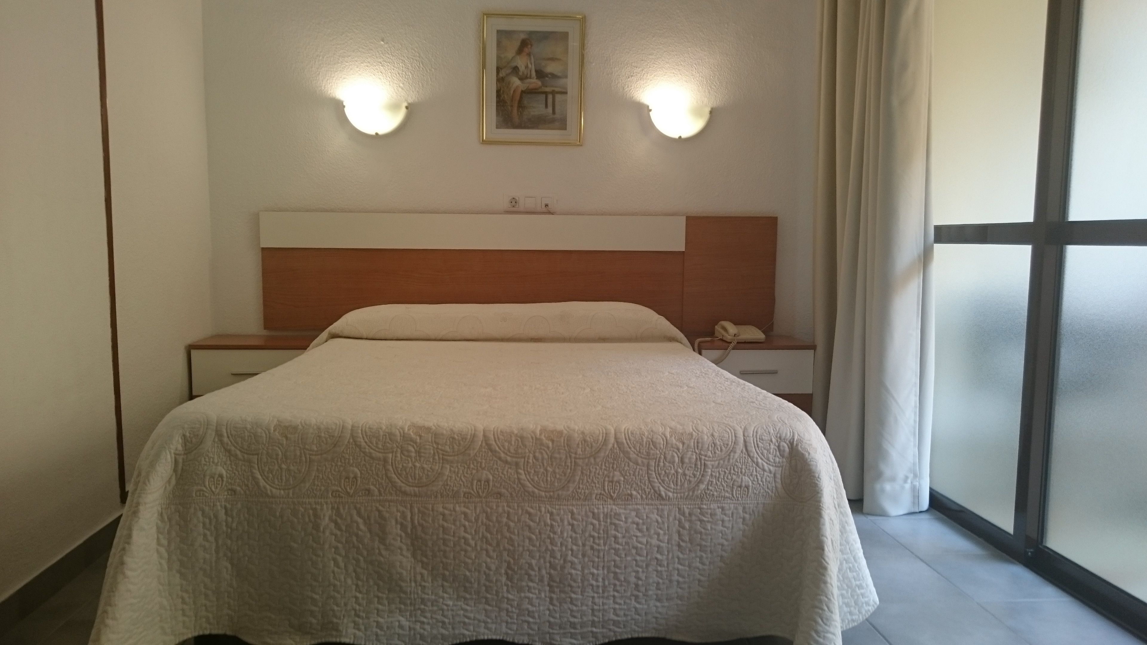 Une chambre d’hôtel avec un grand lit, une tête de lit en bois, deux lampes de chevet, une table de chevet avec un téléphone et une fenêtre avec des rideaux.