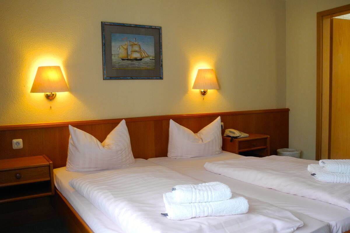 Habitación de hotel con dos camas, cada una con ropa de cama y almohadas blancas, cabeceros de madera, lámparas de noche, un cuadro en la pared y un teléfono en la mesita de noche.