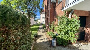 A narrow brick pathway flanked by lush greenery and a building with a visible window and door. - Hotel Pension Kühne (Boltenhagen)